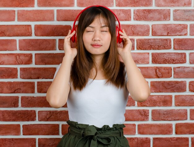 Ritratto di giovane donna asiatica attraente in t-shirt bianca e gonna verde in piedi indossando cuffie rosse ascoltando musica e sorridente con sfondo muro di mattoni arancione.