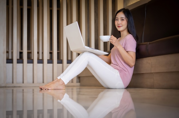 Ritratto di giovane donna asiatica che si siede all'interno in salotto a bere il caffè utilizzando il computer portatile. Lavora da casa concetto.