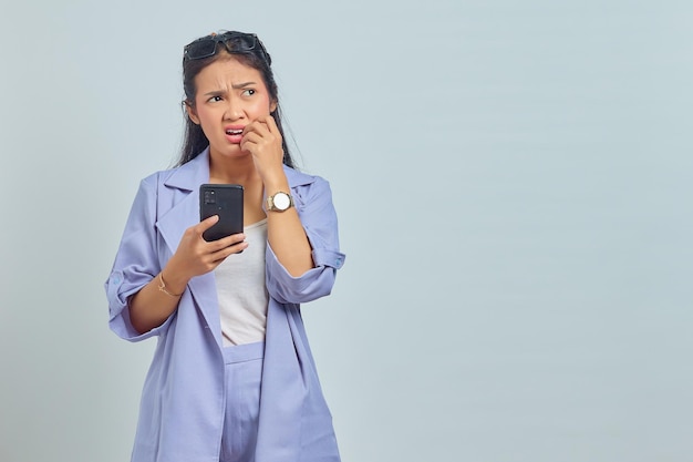 Ritratto di giovane donna asiatica che utilizza il telefono cellulare che sembra stressato e nervoso con la mano in bocca che morde il chiodo su sfondo bianco