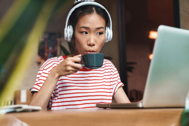 Ritratto di giovane donna asiatica con le cuffie che beve caffè mentre si utilizza il computer portatile nella caffetteria al chiuso