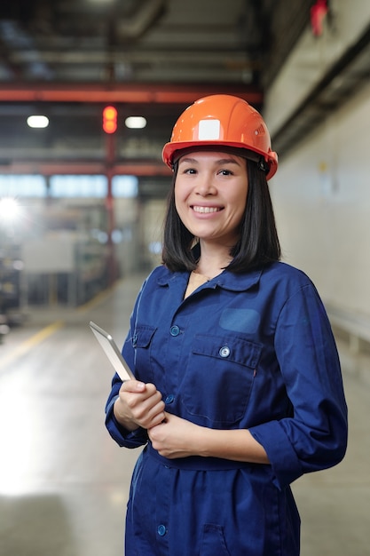 Ritratto di giovane donna asiatica positiva in abbigliamento da lavoro blu e hardhat che tiene compressa in negozio industriale