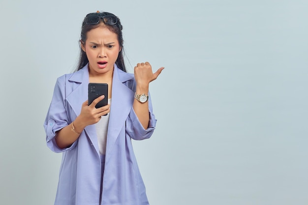 Ritratto di giovane donna asiatica sorpresa che utilizza il telefono cellulare e punta a copiare lo spazio isolato su sfondo bianco