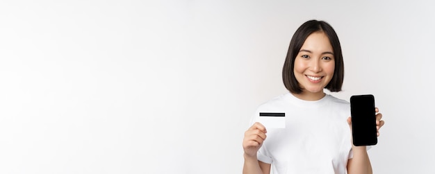 Ritratto di giovane donna asiatica sorridente che mostra lo schermo del telefono cellulare e la carta di credito in piedi sopra il briciolo