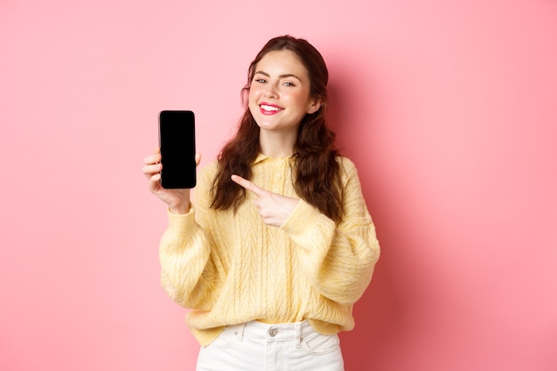 Ritratto di giovane donna attraente dimostra promo sul telefono, puntando il dito sullo smartphone e sorridente, in piedi contro il muro rosa.