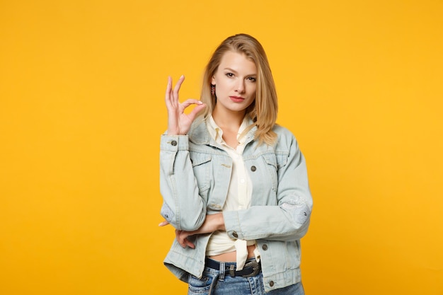 Ritratto di giovane donna attraente e sicura di sé in abiti casual in denim che guarda la fotocamera, mostrando il gesto OK isolato su sfondo giallo arancione in studio. Concetto di stile di vita delle persone. Mock up spazio di copia.