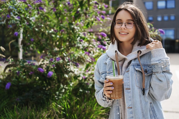 Ritratto di giovane donna attraente in piedi sulla strada vicino a cespugli verdi che bevono caffè preferito ghiaccio latte da asporto fantastico caffè locale sorridente fotocamera soddisfatta camminare all'aperto felice