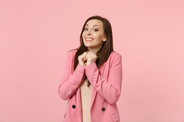 Ritratto di giovane donna attraente piuttosto sorridente in giacca che stringe i pugni vicino al viso isolato su sfondo rosa pastello in studio. Persone sincere emozioni, concetto di stile di vita. Mock up spazio di copia.