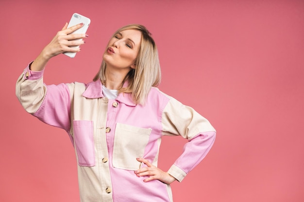 Ritratto di giovane donna bella bionda sorridente allegra eccitata in camicia casual che fa foto selfie isolata su sfondo rosa. Utilizzo del cellulare.