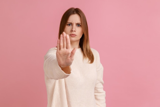 Ritratto di giovane donna bionda adulta seria e seria in piedi con il gesto delle mani di arresto e attenzione a non toccare indossando un maglione bianco Colpo in studio al coperto isolato su sfondo rosa
