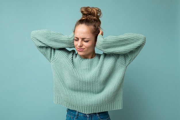 Ritratto di giovane donna bionda attraente insoddisfatta triste emotiva con emozioni sincere che indossa la maglia blu casual isolata su sfondo blu con spazio vuoto e copre le orecchie