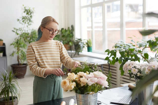 Ritratto di giovane donna bionda che dispone fiori e crea mazzi di fiori mentre lavora in un laboratorio di fioristi verdi, copia spazio
