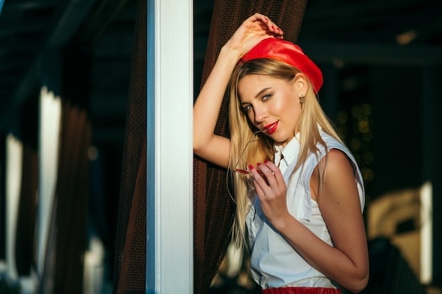 Ritratto di giovane donna bionda in berretto rosso e pantaloncini rossi in pelle in posa all'aperto Trucco luminoso