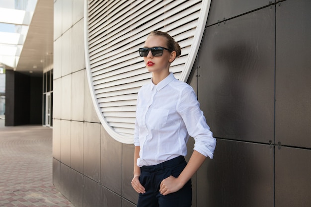 Ritratto di giovane donna bionda in occhiali da sole che posano contro la parete dell'edificio all'aperto