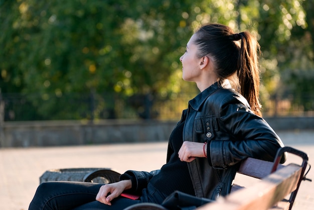 Ritratto di giovane donna bruna all'aperto. Riposo su una panchina nel parco. Vista laterale.