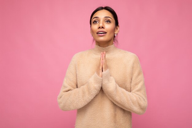 Ritratto di giovane donna bruna attraente con emozioni sincere che indossa pullover beige isolato su sfondo rosa con spazio copia e tenendo la mano nel gesto di preghiera credendo nella buona fortuna