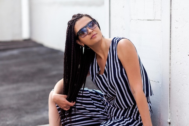 Ritratto di giovane donna bruna con l'acconciatura afro americana nera dreadlocks in abito a righe e occhiali da sole seduto in posa tenendo i capelli e guardando il modello di moda della fotocamera girato