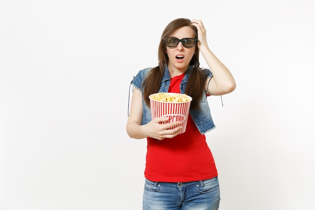 Ritratto di giovane donna bruna scioccata irritata in occhiali 3d guardando film, tenendo secchio di popcorn e tenendo la mano vicino alla testa isolata su sfondo bianco. Emozioni nel concetto di cinema.