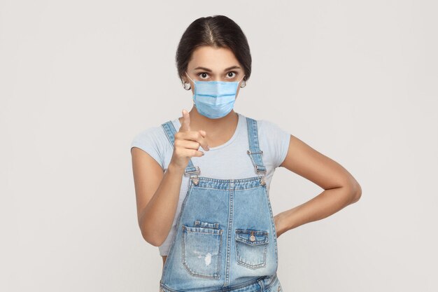 Ritratto di giovane donna bruna seria con maschera medica chirurgica in tuta di jeans in piedi guardando la macchina fotografica e avvertendo o rimproverando. studio al coperto colpo isolato su sfondo grigio.