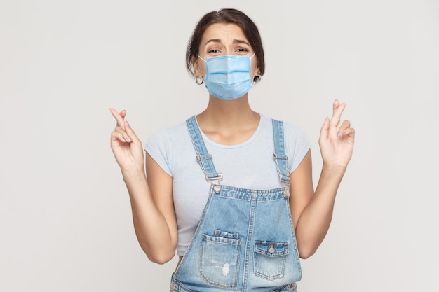 Ritratto di giovane donna bruna speranzosa con maschera medica chirurgica in tuta di jeans in piedi con le braccia incrociate, guardando la telecamera con un desiderio di preoccupazione. studio al coperto colpo isolato su sfondo grigio.