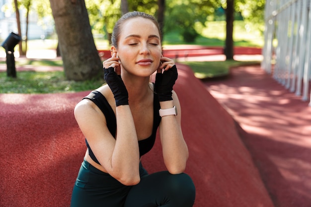 Ritratto di giovane donna calma che indossa tuta da ginnastica e guanti che riposa dopo l'allenamento con barra di metallo orizzontale sul campo sportivo nel parco verde