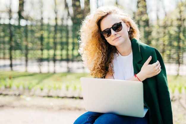 Ritratto di giovane donna carina con capelli biondi ricci che indossa occhiali da sole maglietta bianca e giacca verde che tiene il laptop sulle ginocchia che lavora al suo progetto futuro isolato su sfondo verde natura