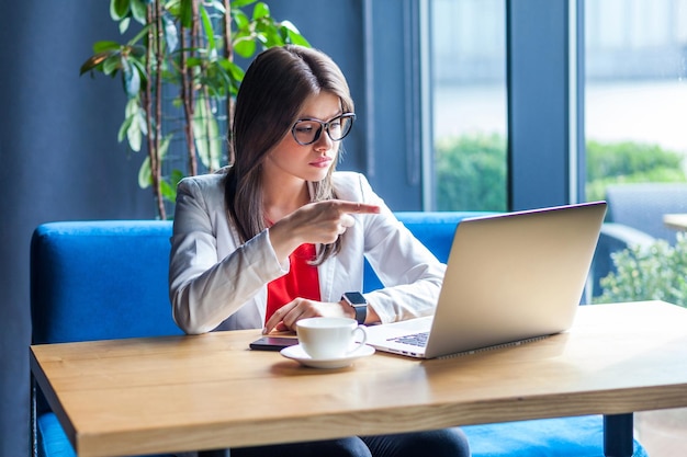 Ritratto di giovane donna castana bella ed elegante seria con gli occhiali seduto a guardare lo schermo del suo laptop durante la videochiamata e accusando o incolpando lo studio al coperto sparato sullo sfondo dell'ufficio del caffè