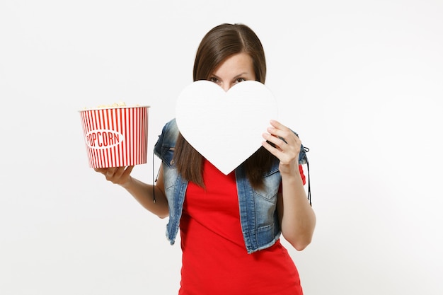 Ritratto di giovane donna castana in abiti casual che guarda film, tiene in mano un secchio di popcorn e copre il viso con cuore bianco con spazio copia isolato su sfondo bianco. Emozioni al cinema.