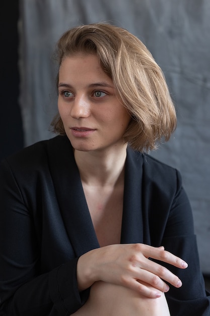 Ritratto di giovane donna caucasica con i capelli corti in posa in giacca di abito nero, seduto davanti al tessuto grigio. bella ragazza sorride. pose femminili attraenti dai capelli corti in studio