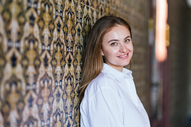 Ritratto di giovane donna caucasica sorridente in camicia bianca appoggiata al muro piastrellato