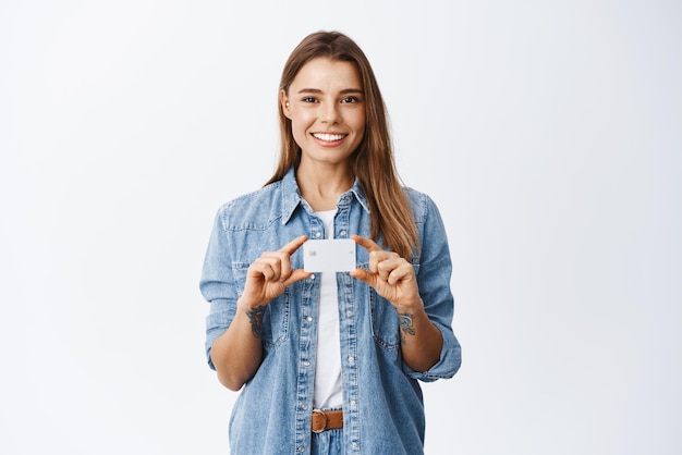 Ritratto di giovane donna che consiglia banca mostrando carta di credito in plastica sul petto e sorridente dando consigli per il deposito in piedi su sfondo bianco