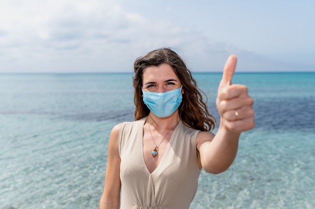 Ritratto di giovane donna che indossa una maschera protettiva che mostra pollice in su. Bellissimo sfondo con acqua limpida blu