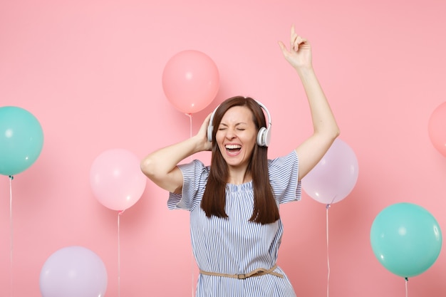 Ritratto di giovane donna che ride con gli occhi chiusi con le cuffie che indossano abiti blu ascoltando musica ballando alzando le mani su sfondo rosa con mongolfiera colorata. Concetto di festa di compleanno.