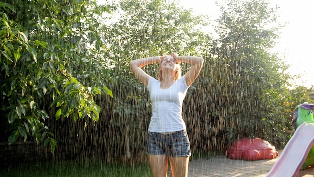 Ritratto di giovane donna che ride felice con i capelli lunghi in vestiti bagnati che ballano sotto la pioggia calda in giardino. La famiglia gioca e si diverte all'aperto in estate