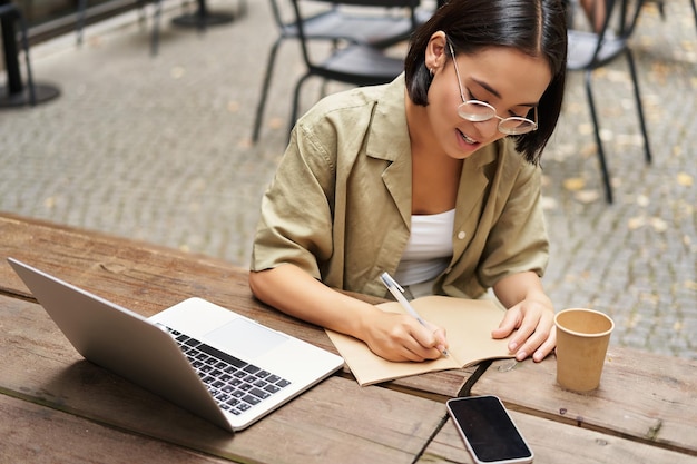 Ritratto di giovane donna che studia online seduta con il computer portatile che scrive prendendo appunti e guardando