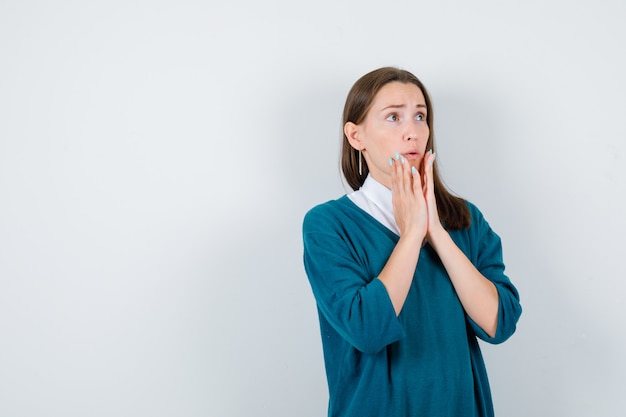 Ritratto di giovane donna che tiene le mani vicino alla bocca, alzando lo sguardo in un maglione sopra la camicia bianca e guardando la vista frontale spaventata