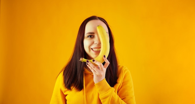 Ritratto di giovane donna con banana su sfondo giallo Femmina divertente in felpa con cappuccio gialla in posa con frutta