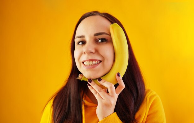 Ritratto di giovane donna con banana su sfondo giallo Femmina divertente in felpa con cappuccio gialla in posa con frutta