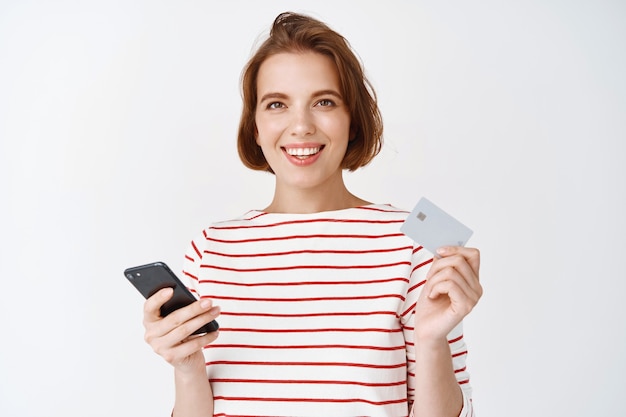 Ritratto di giovane donna con i capelli corti, shopping online, mostrando carta di credito in plastica e pagando su smartphone, sorridente, muro bianco