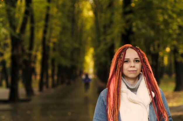 Ritratto di giovane donna con i dreadlocks allo zenzero che posano nel parco forestale Bella donna con acconciatura brillante in una calda giacca di jeans a piedi