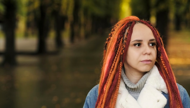 Ritratto di giovane donna con i dreadlocks allo zenzero che posano nel parco forestale Bella donna con acconciatura brillante in una calda giacca di jeans a piedi