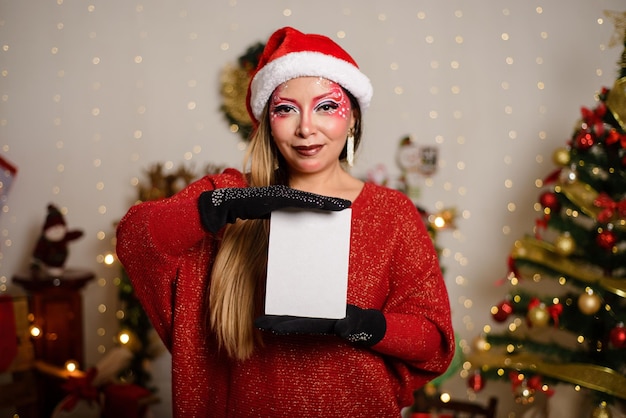 Ritratto di giovane donna con il trucco di Natale che indossa il cappello di Babbo Natale in un set di Natale in possesso di una cartolina vuota Concentrarsi sulla cartolina