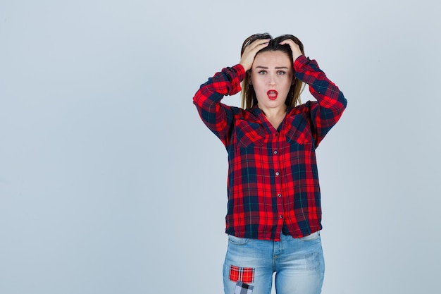 Ritratto di giovane donna con le mani sulla testa, bocca aperta in camicia a quadri, jeans e vista frontale scioccata