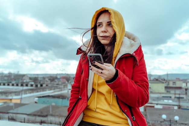 Ritratto di giovane donna con telefono cellulare sul tetto dell'edificio Bruna con felpa con cappuccio gialla e giacca rossa che naviga sullo smartphone con tempo nuvoloso