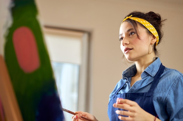 Ritratto di giovane donna creativa in grembiule che guarda il suo lavoro mentre lavora alla pittura astratta a