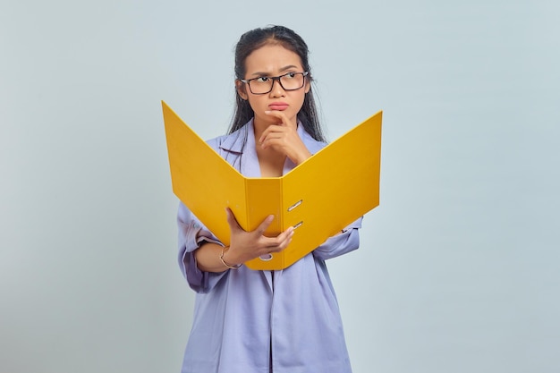 Ritratto di giovane donna d'affari asiatica seria che tiene il mento, contemplando qualcosa mentre tiene la cartella dei documenti isolata su sfondo viola