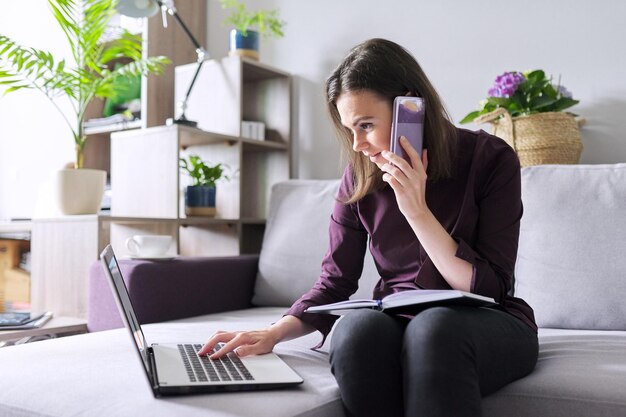 Ritratto di giovane donna d'affari che lavora a casa Donna seduta sul divano con il portatile parlando al telefono prendendo appunti in un notebook aziendale