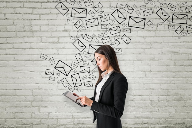 Ritratto di giovane donna d'affari europea attraente che utilizza il tablet con icone e-mail disegnate creative su sfondo bianco muro di mattoni Comunicazione tecnologica aziendale e concetto di marketing via e-mail