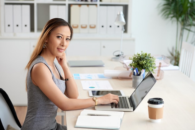 Ritratto di giovane donna dell'ufficio vietnamita di contenuti che utilizza il computer portatile a tavola con una tazza di caffè e un organizer