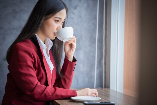 Ritratto di giovane donna di affari asiatica che si siede all'interno nel caffè bevente del caffè con lo Smart Phone. Concetto di successo aziendale.
