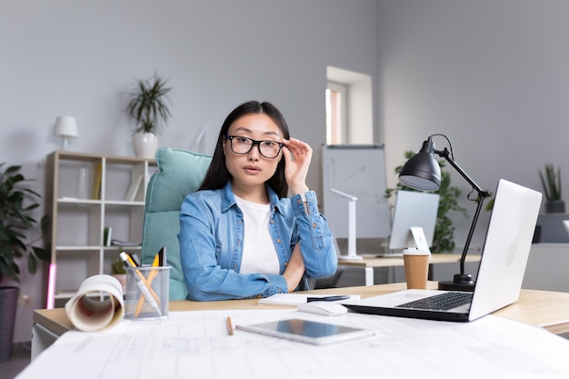 Ritratto di giovane donna di design in un ufficio moderno al lavoro Donna asiatica di successo con gli occhiali che guarda l'obbiettivo